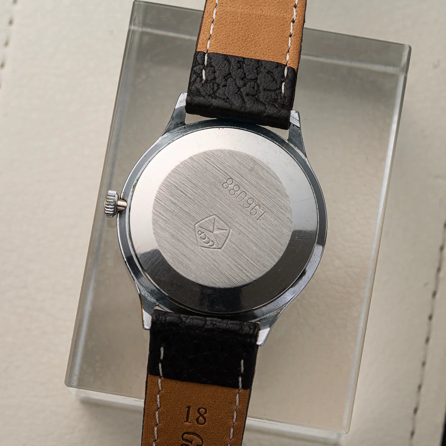 A close-up of a wristwatch Slava Roman Numerals shown from the back, featuring a brushed silver metal case and a brown leather strap with black textured stitching and a black croc-embossed section near the top. The watch is displayed on a clear acrylic stand against a light beige textured background.