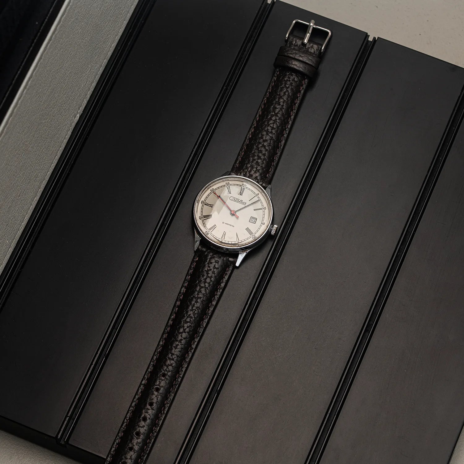 A vintage-style wristwatch Slava Roman Numerals with a silver-toned oval case, white dial, and textured dark brown leather strap, displayed diagonally on a black presentation box.