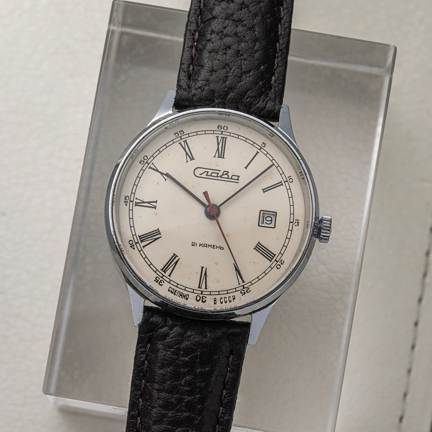 A vintage-style analog wristwatch Slava Roman Numerals with a silver-toned round case, cream dial, black Roman numerals, and a black textured leather strap, displayed upright on a clear acrylic stand.