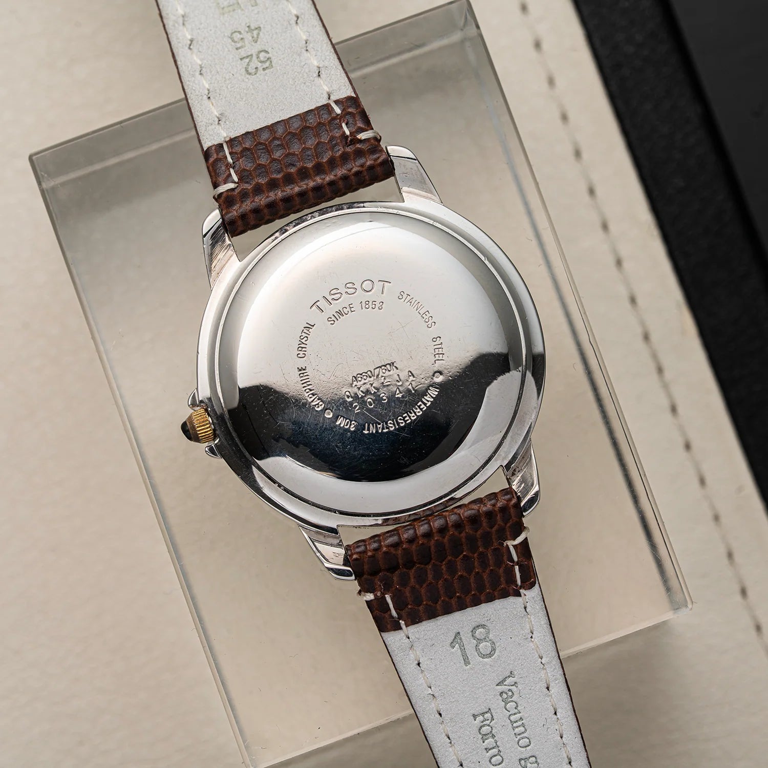 Close-up of the back of a Tissot Seastar ETA 2846 wristwatch with a silver stainless steel case and a brown textured leather strap, resting on a clear display block.