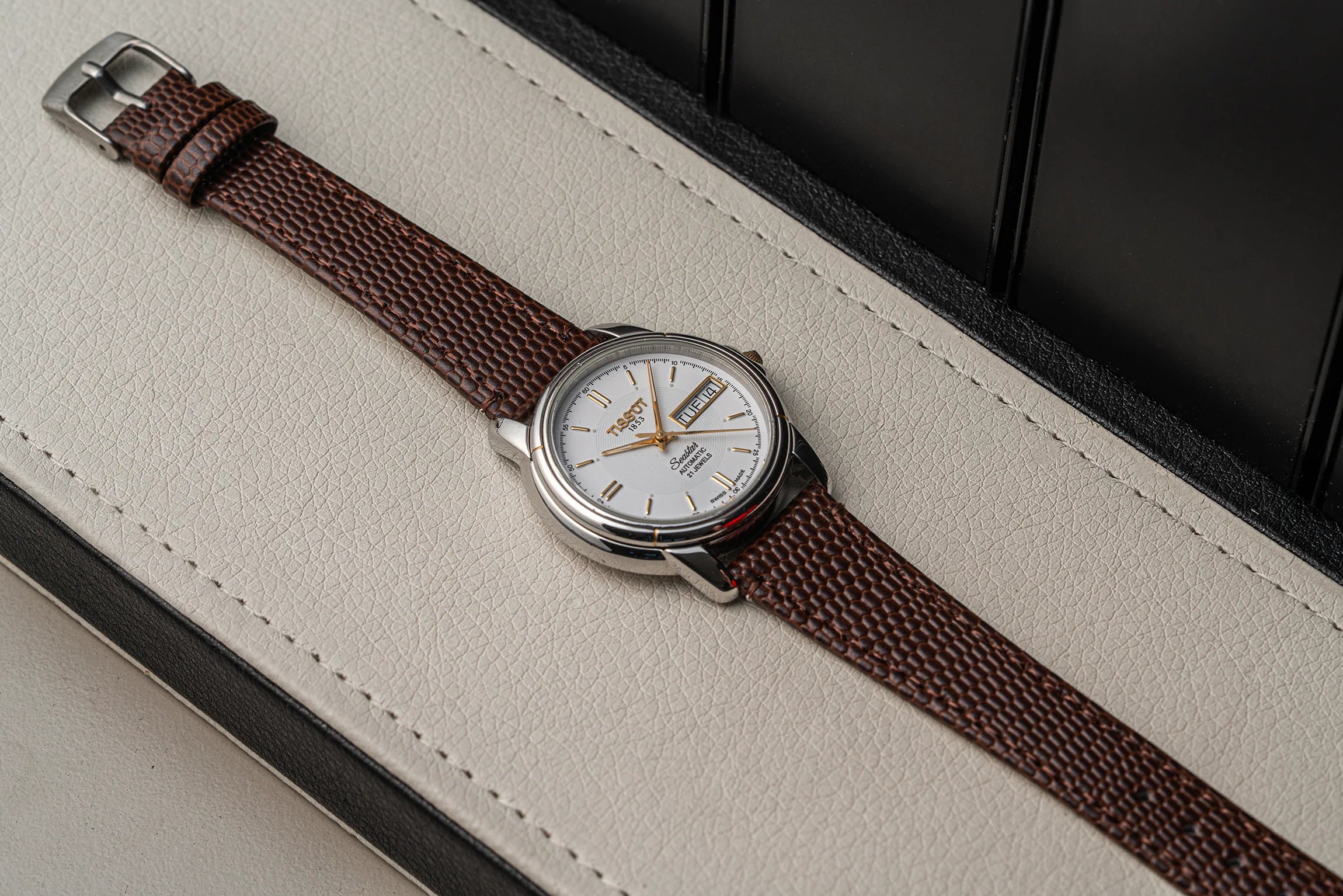 A wristwatch Tissot Seastar ETA 2846 with a brown woven strap and a silver-toned round case, resting diagonally on a cream-colored textured leather surface.