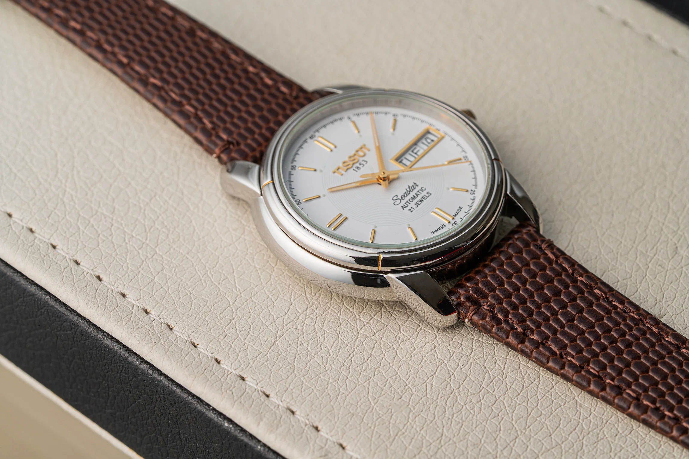 A close-up of a Tissot Seastar ETA 2846 watch with a brown textured strap and a silver-toned case, resting on a light beige textured surface.