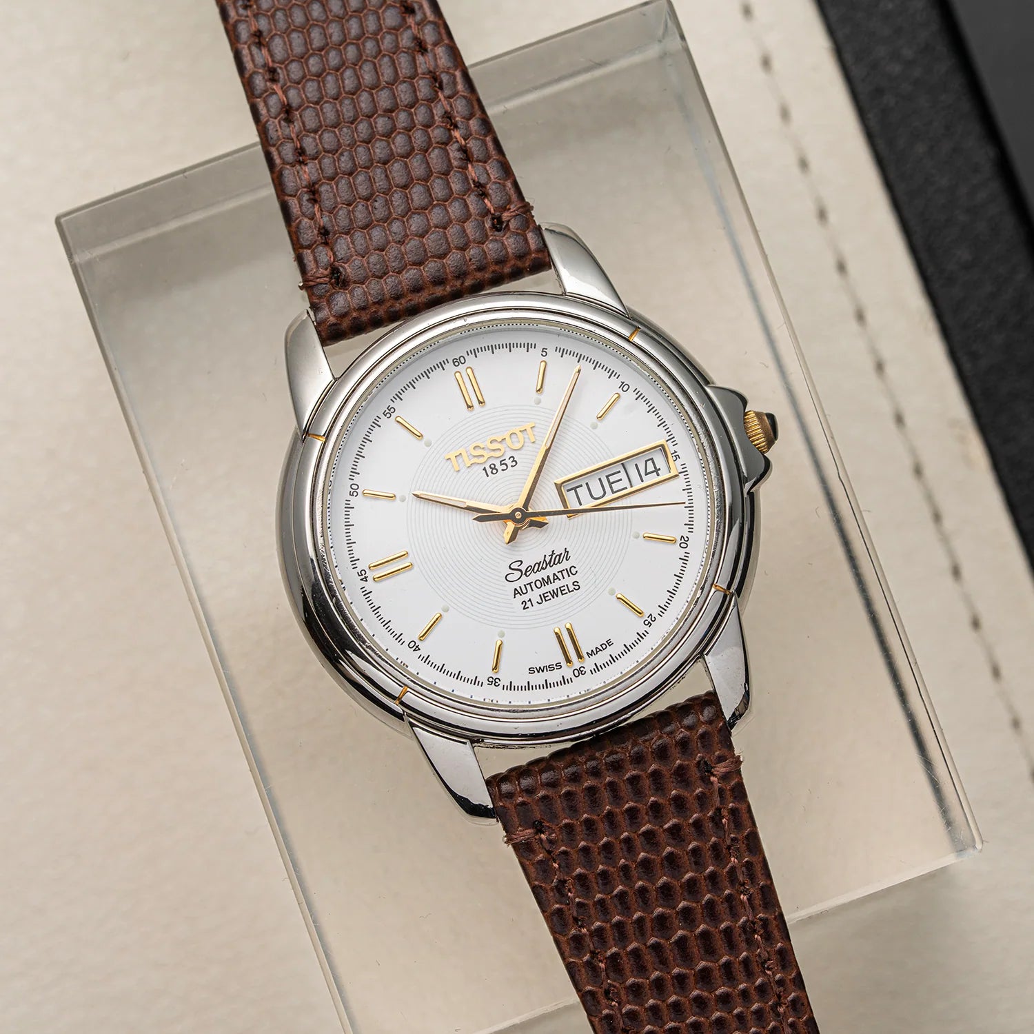 A Tissot Seastar ETA 2846 Automatic watch with a silver stainless steel case, white dial, and textured brown leather strap, displayed on a clear acrylic stand.