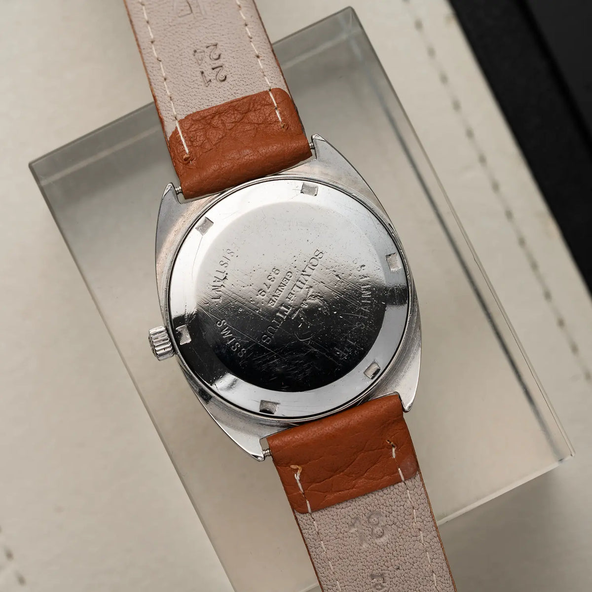 Close-up of the back of a vintage wristwatch Titus Geneve, Ref. 9379, Cal. FHF 96-4, Men’s Manual Wind, 1972 with a silver metal case and a brown leather strap, resting on a clear acrylic block.