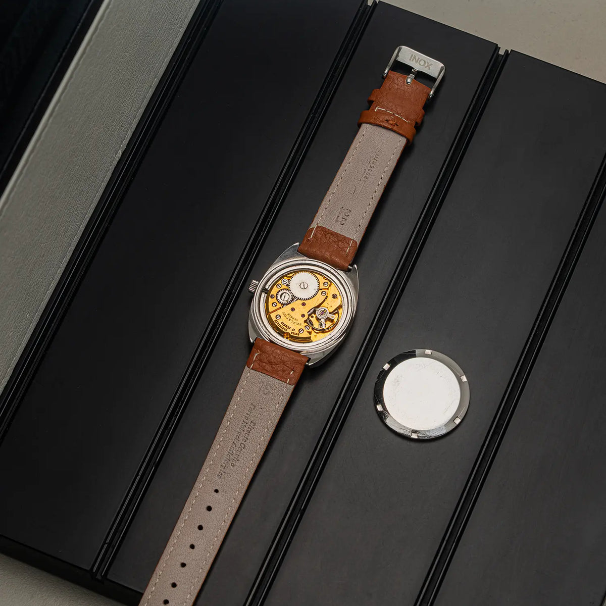 A luxury watch with a silver-toned case, a gold-toned mechanical movement visible through the back, and a light brown leather strap, displayed diagonally on a black presentation box next to its circular metal lid.