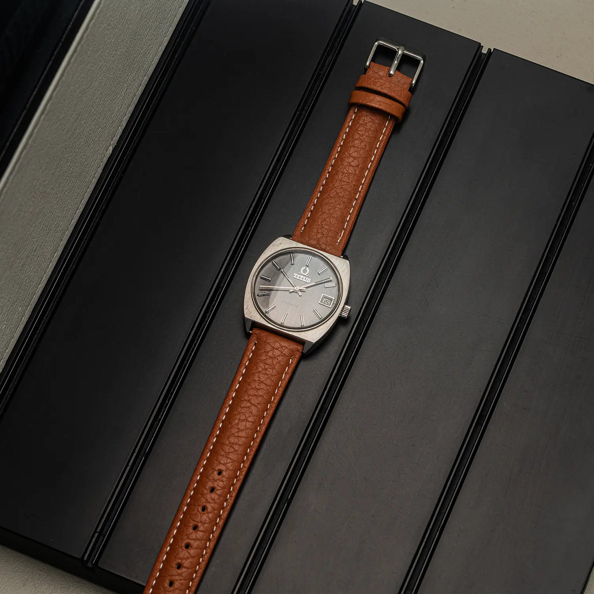 A vintage-style wristwatch Titus Geneve, Ref. 9379, Cal. FHF 96-4, Men’s Manual Wind, 1972 with a silver-toned square case, silver dial, and a brown textured leather strap, displayed diagonally on a black textured surface.