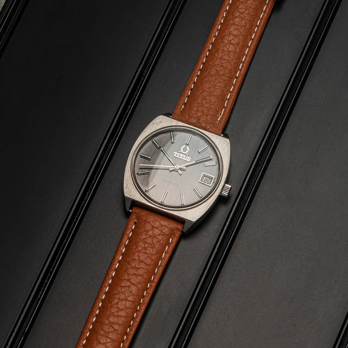 A vintage-style wristwatch Titus Geneve, Ref. 9379, Cal. FHF 96-4, Men’s Manual Wind, 1972 with a silver-toned rectangular case, silver dial, and brown textured leather strap with white stitching, photographed on a dark striped surface.