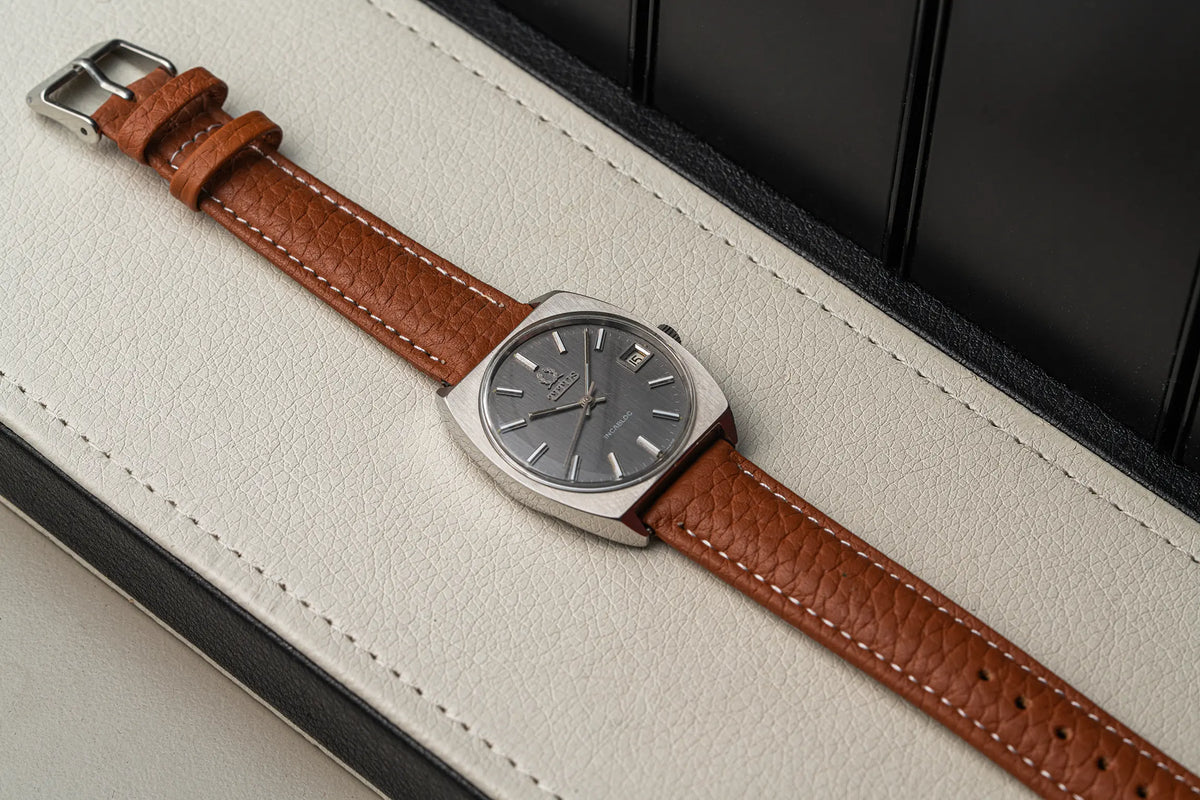 A vintage-style wristwatch Titus Geneve, Ref. 9379, Cal. FHF 96-4, Men’s Manual Wind, 1972 with a brown textured leather strap and a silver-toned rectangular case, shown resting on a cream-colored textured surface with a dark background.
