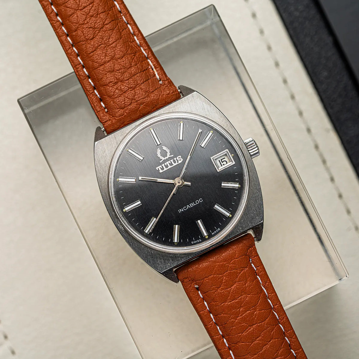 A vintage Titus Geneve, Ref. 9379, Cal. FHF 96-4, Men’s Manual Wind, 1972wristwatch with a round black dial, silver-tone case, and a brown leather strap, displayed on a clear acrylic stand.