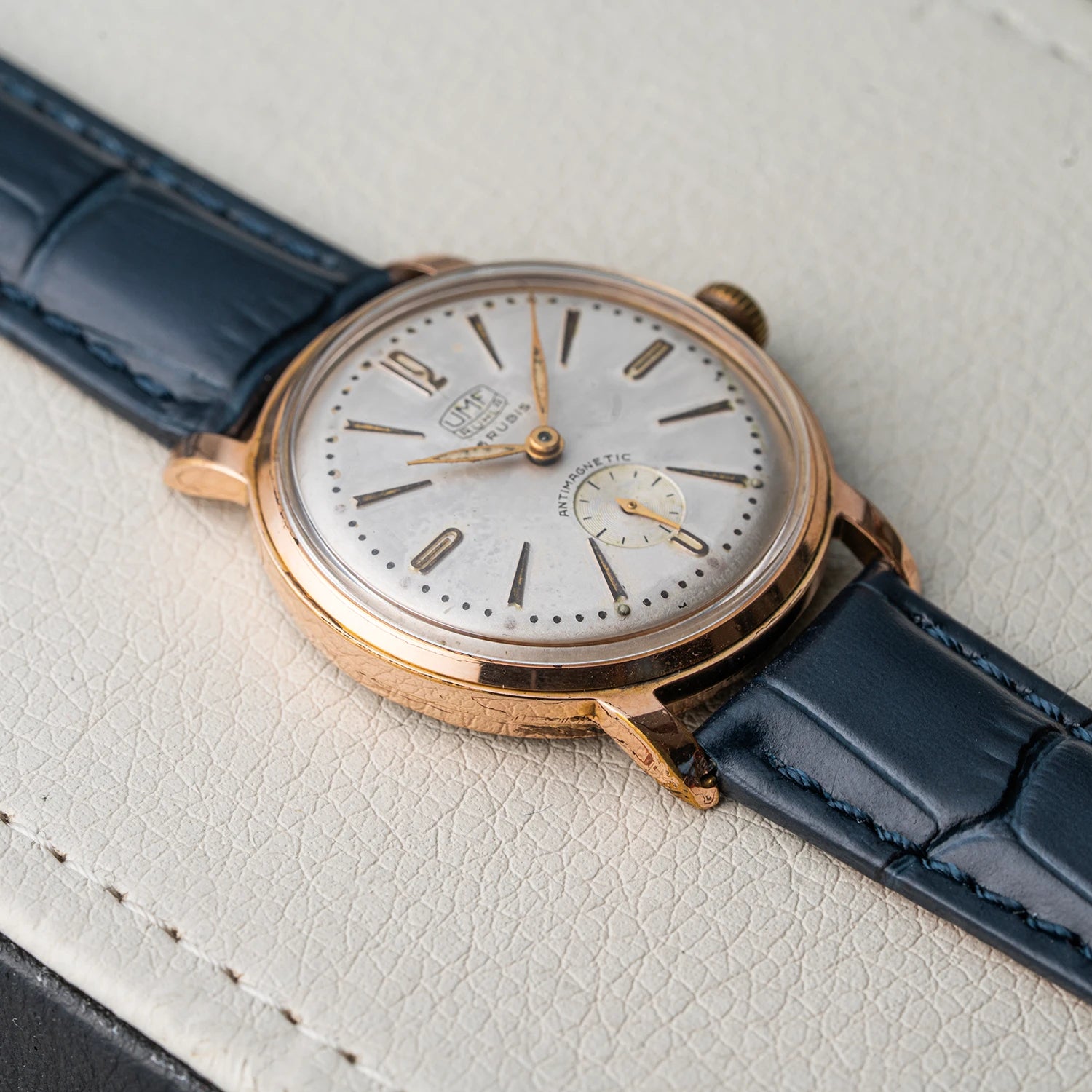 A vintage UMF Ruhla, Cal. UMF 2 wristwatch with a rose gold case, white dial, and blue leather strap, resting on a textured off-white leather surface.