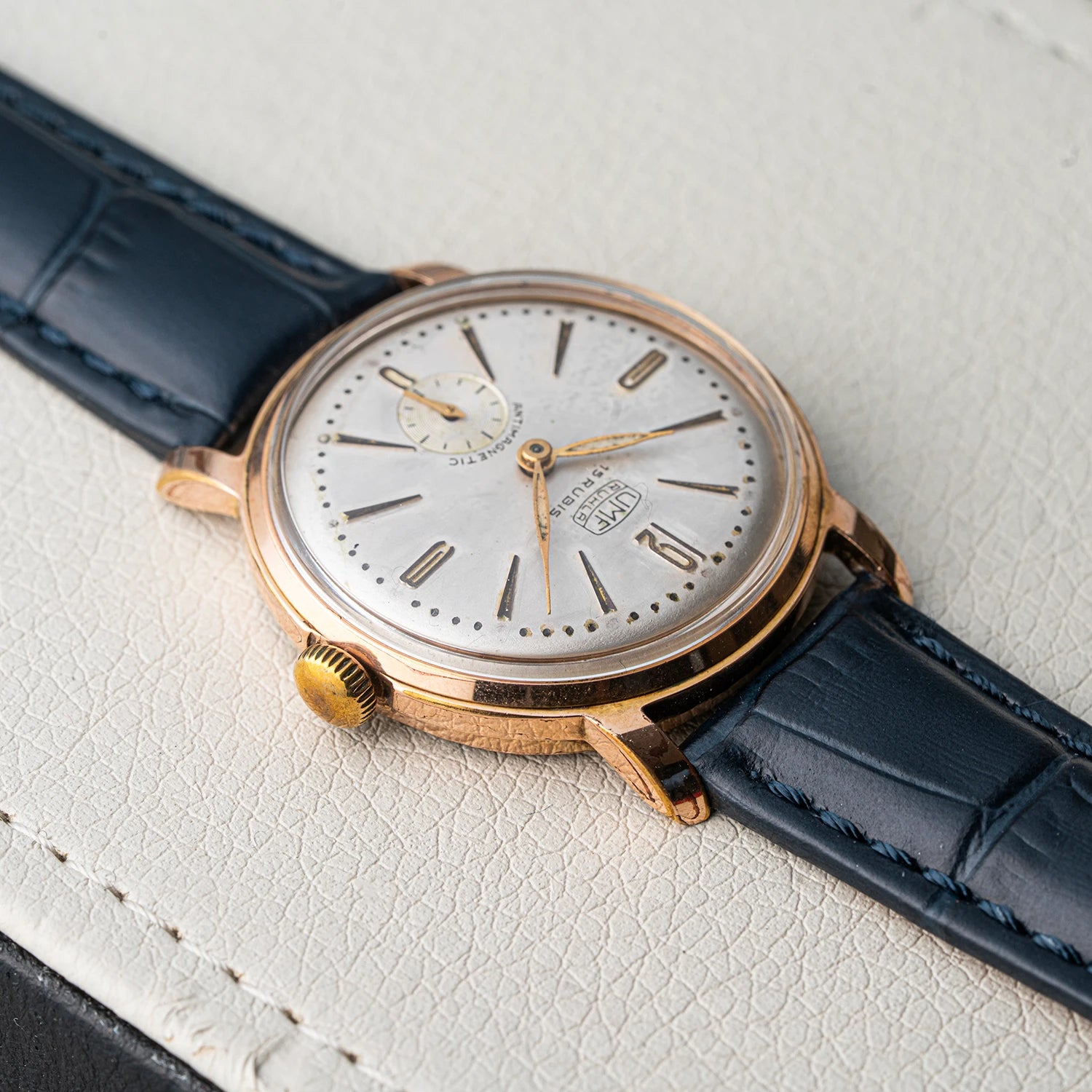 A vintage wristwatch UMF Ruhla, Cal. UMF 2 with a round rose gold case, a silver dial with black stick indices, and a dark blue leather strap, shown resting on a textured off-white leather surface.
