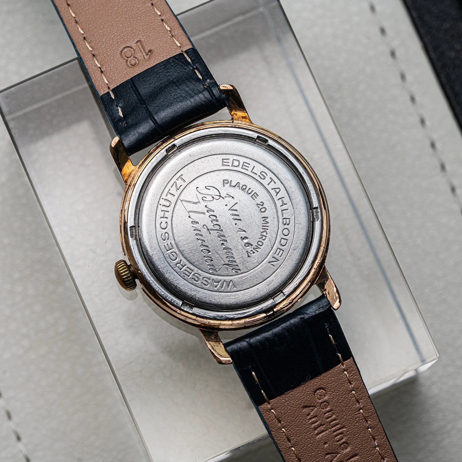 Close-up of the back of a vintage wristwatch with a rose gold case and a dark blue leather strap, resting on a clear display box.