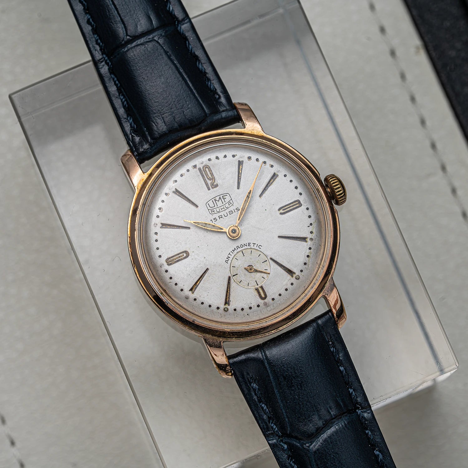 A vintage UMF Ruhla, Cal. UMF 2 wristwatch with a rose gold case, white dial, and dark blue leather strap, resting on a clear display box.