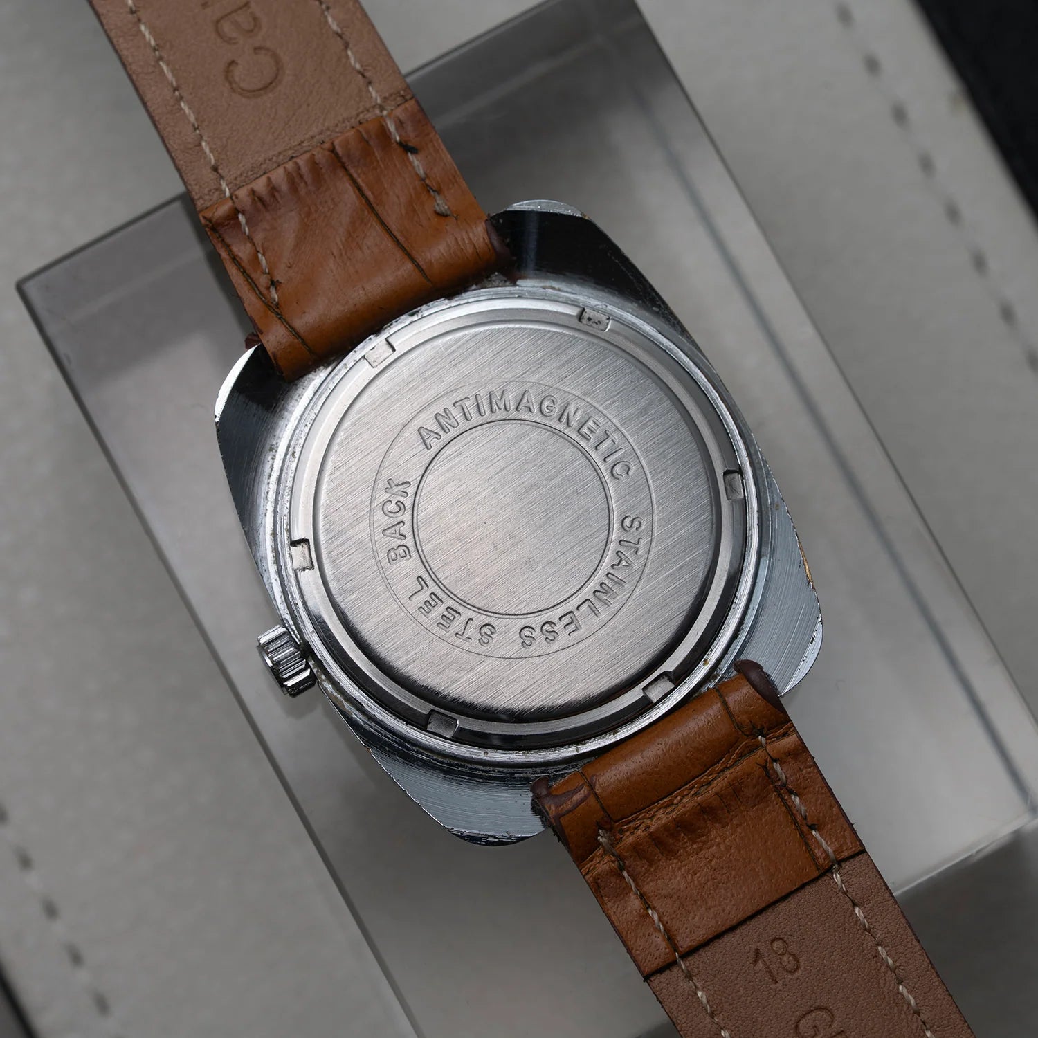 Close-up of the back of a wristwatch with a round brushed stainless steel case and a brown leather strap, resting on a light gray surface.