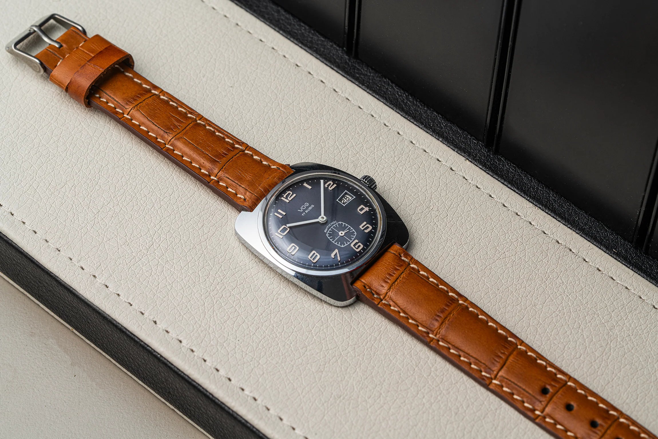 A wristwatch Vog 17 with a brown leather strap and a silver-toned rectangular case, photographed diagonally on a cream-colored textured surface with a dark background.