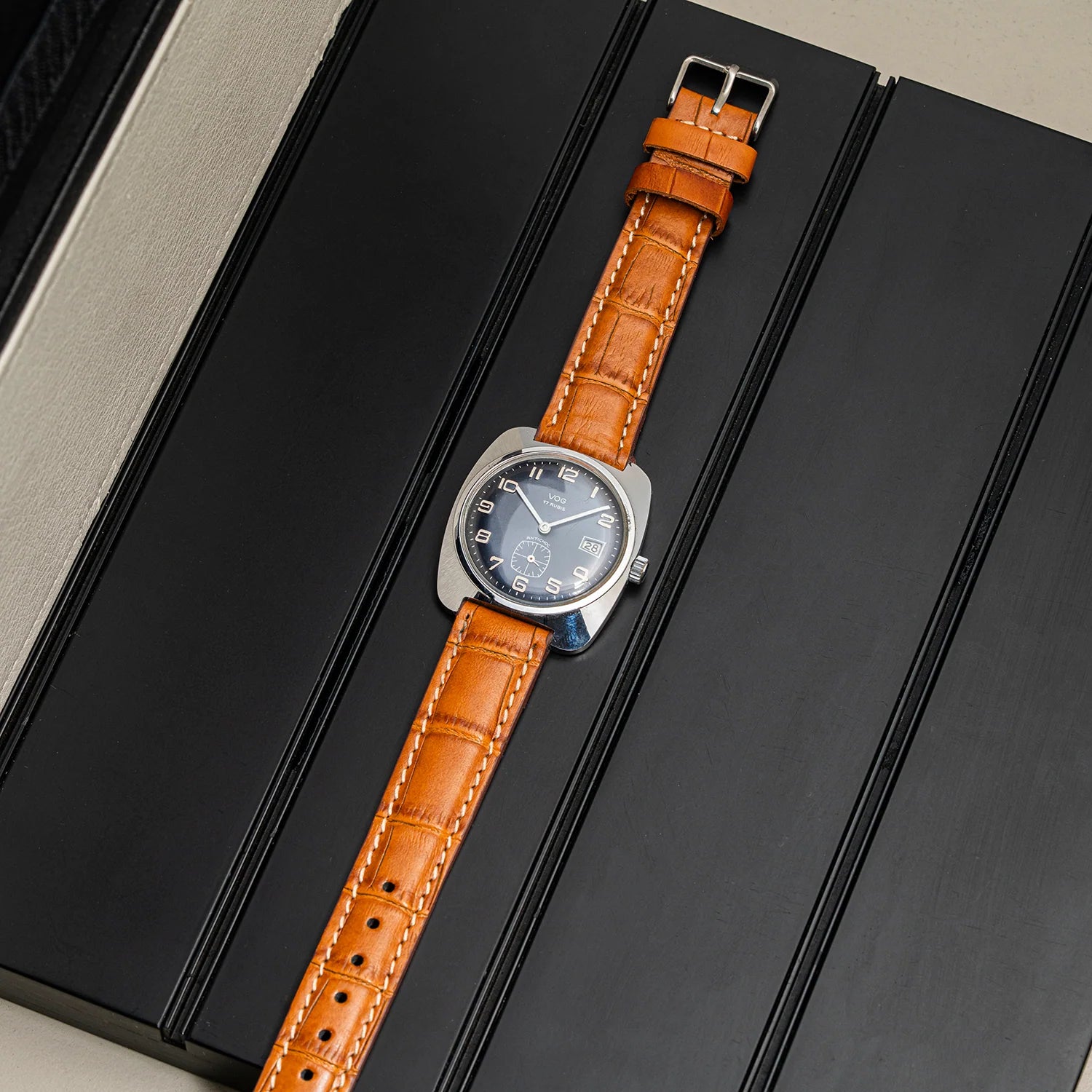 A wristwatch Vog 17 with a round silver-tone case and a tan leather strap, laid diagonally on a black textured surface.