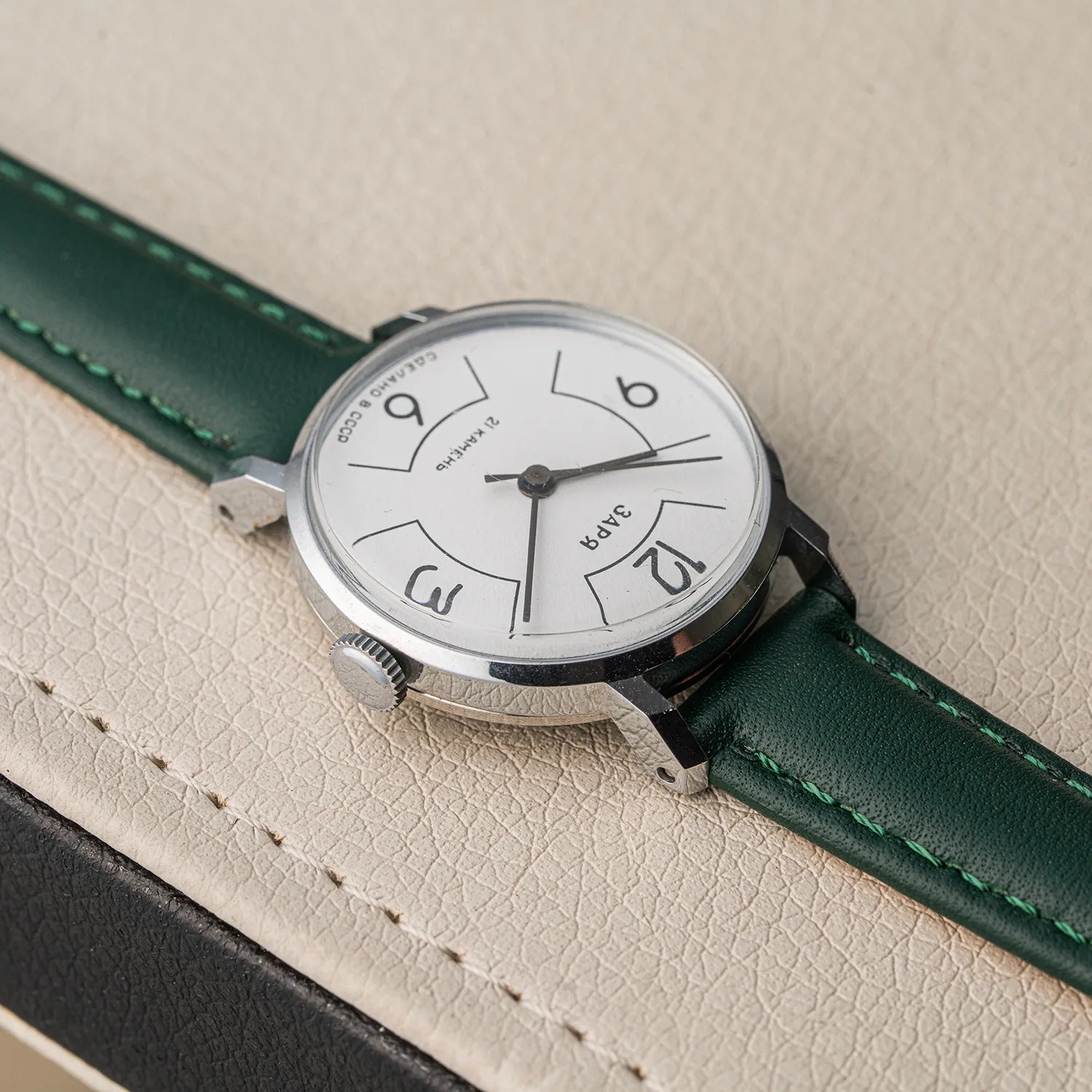 A close-up of a wristwatch Zaria Z2009B with a silver-toned case, white dial, and dark green leather strap with matching green stitching, resting on a textured beige surface.