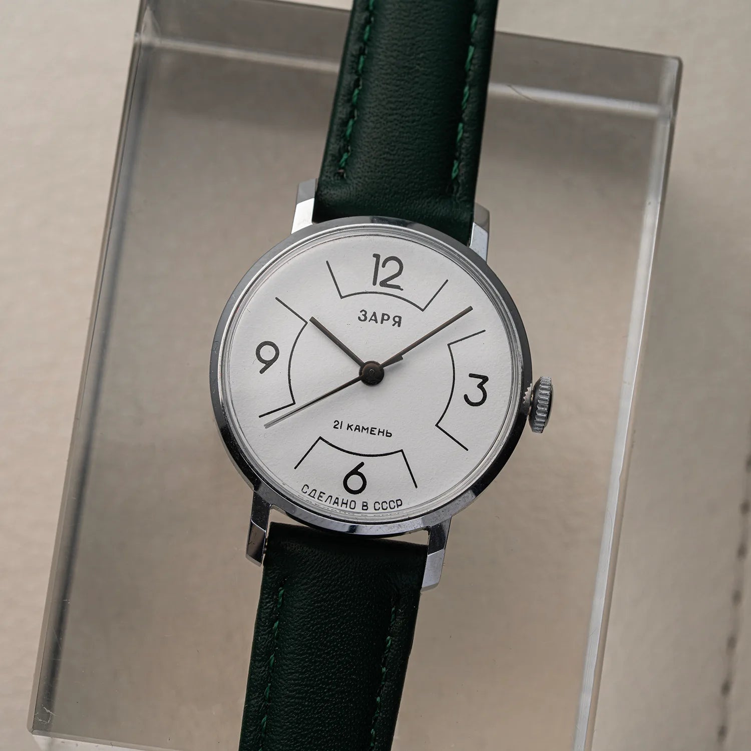 A vintage-style women's wristwatch Zaria Z2009B with a white dial, black Arabic numerals, and a dark green leather strap, displayed inside a clear acrylic case on a beige surface.