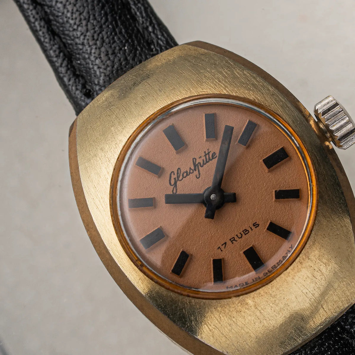Close-up of a vintage Glashutte 17 Rubis wristwatch with a gold-tone case, tan dial, black hands and hour markers, and a black textured leather strap.