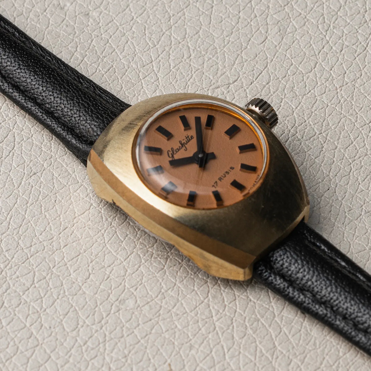A vintage women's wristwatch with a gold-tone case, a tan dial featuring black hands and hour markers, and a black leather strap, resting on a textured light beige surface.