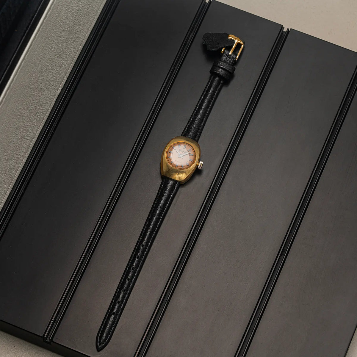 A vintage-style wristwatch with a gold-tone oval case and a black leather strap, displayed diagonally on a black presentation box.
