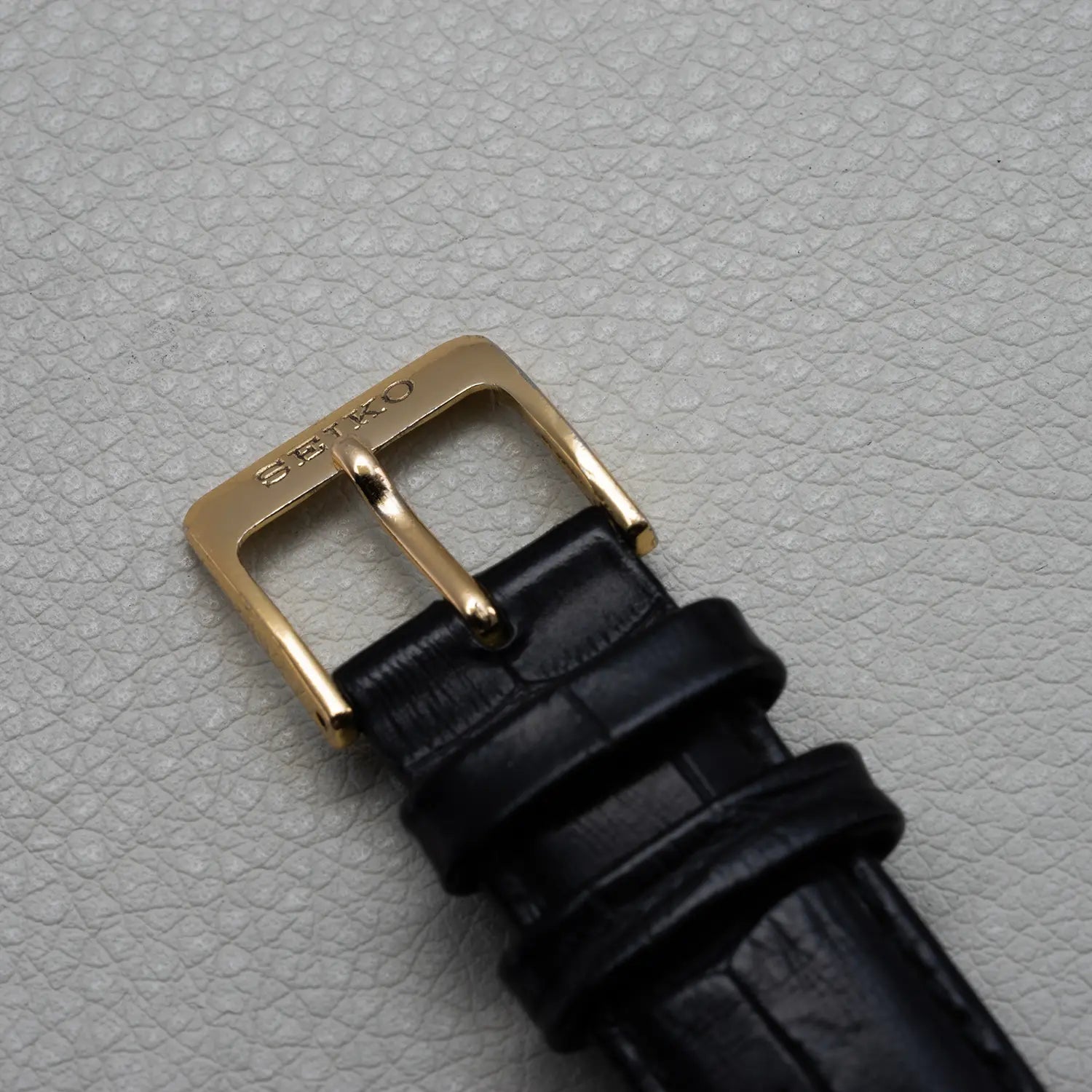 Black leather watch strap with gold buckle on a light gray textured background