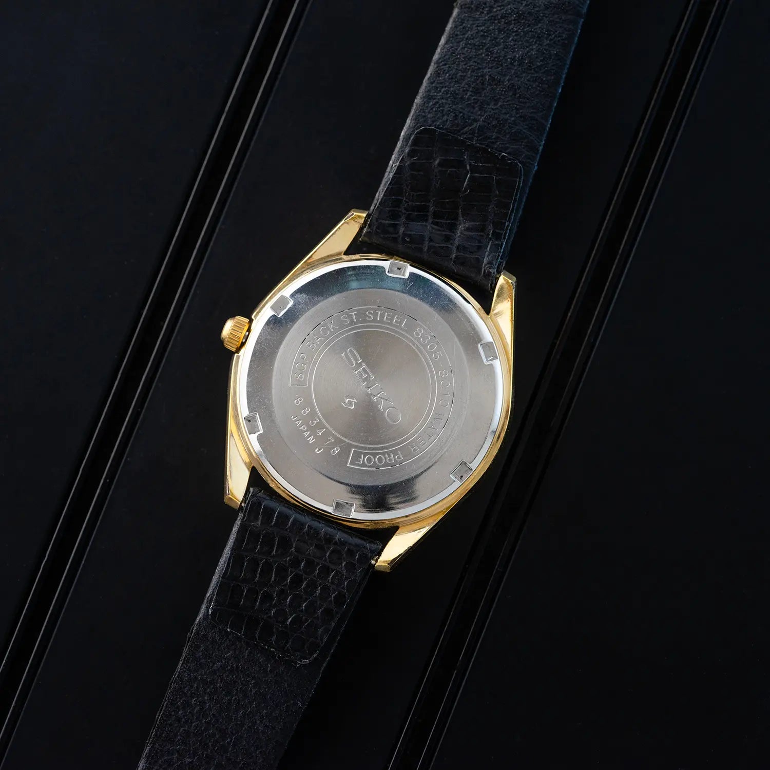 Wristwatch Seiko 8305 8010 case back with black strap and gold case on a black background
