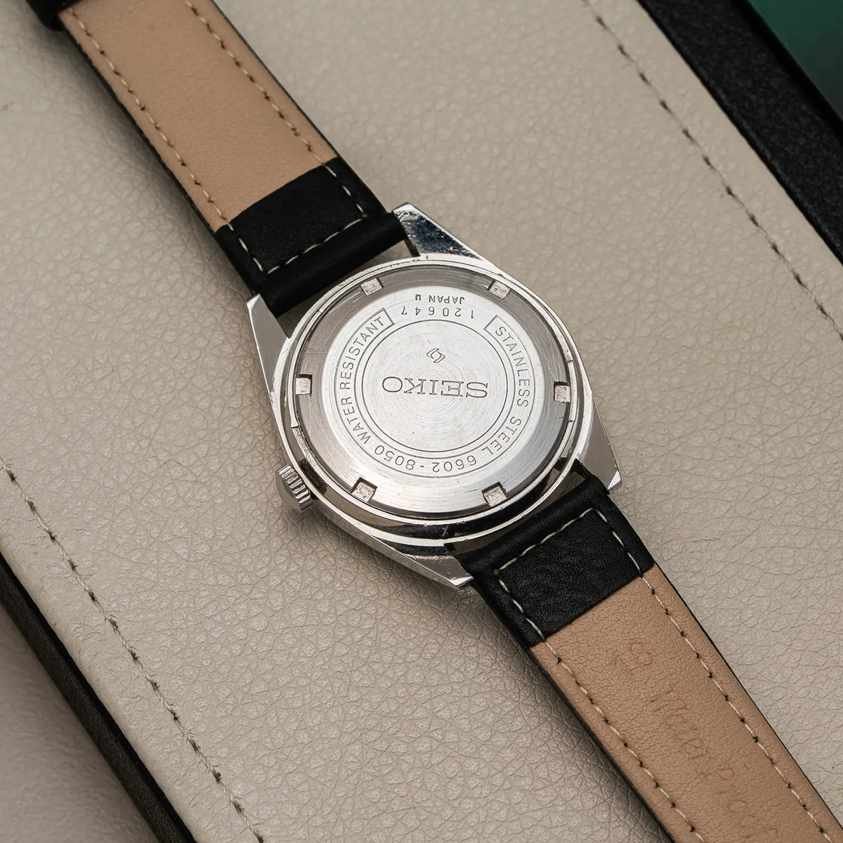 Close-up of the back of a wristwatch Seiko, Ref. 6602-8050, Cal. 6602, Men’s Manual Wind, 1971 with a silver stainless steel case and a tan leather strap with black stitching, resting on a light beige textured surface.