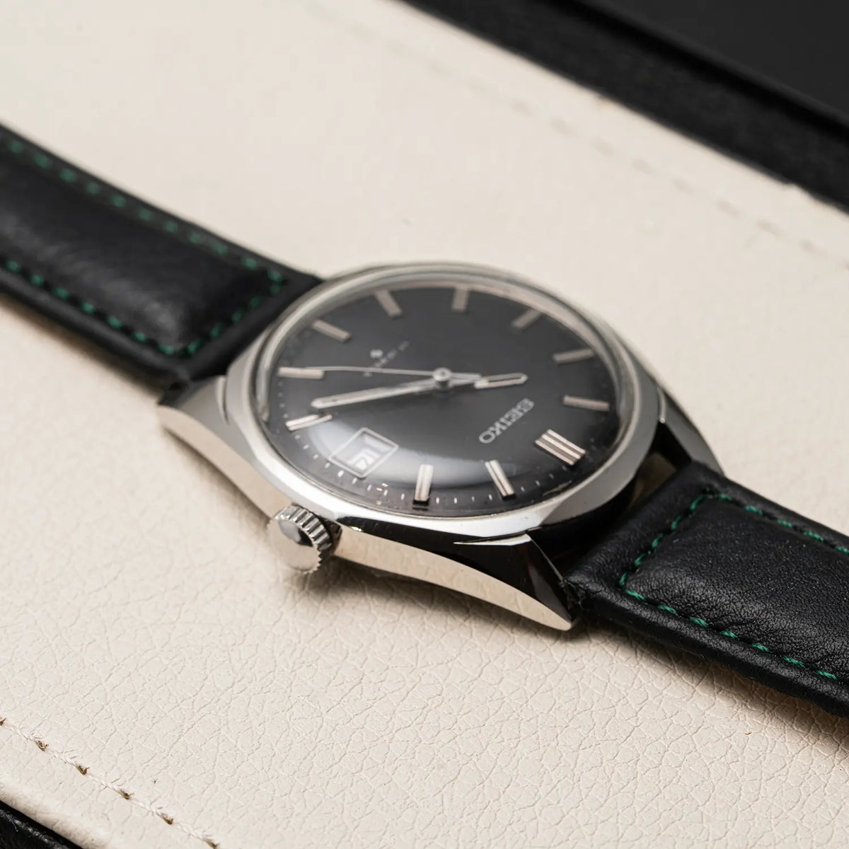 A close-up of a Seiko, Ref. 6602-8050, Cal. 6602, Men’s Manual Wind, 1971 wristwatch with a black dial and black leather strap with green stitching, resting on a textured cream-colored surface.