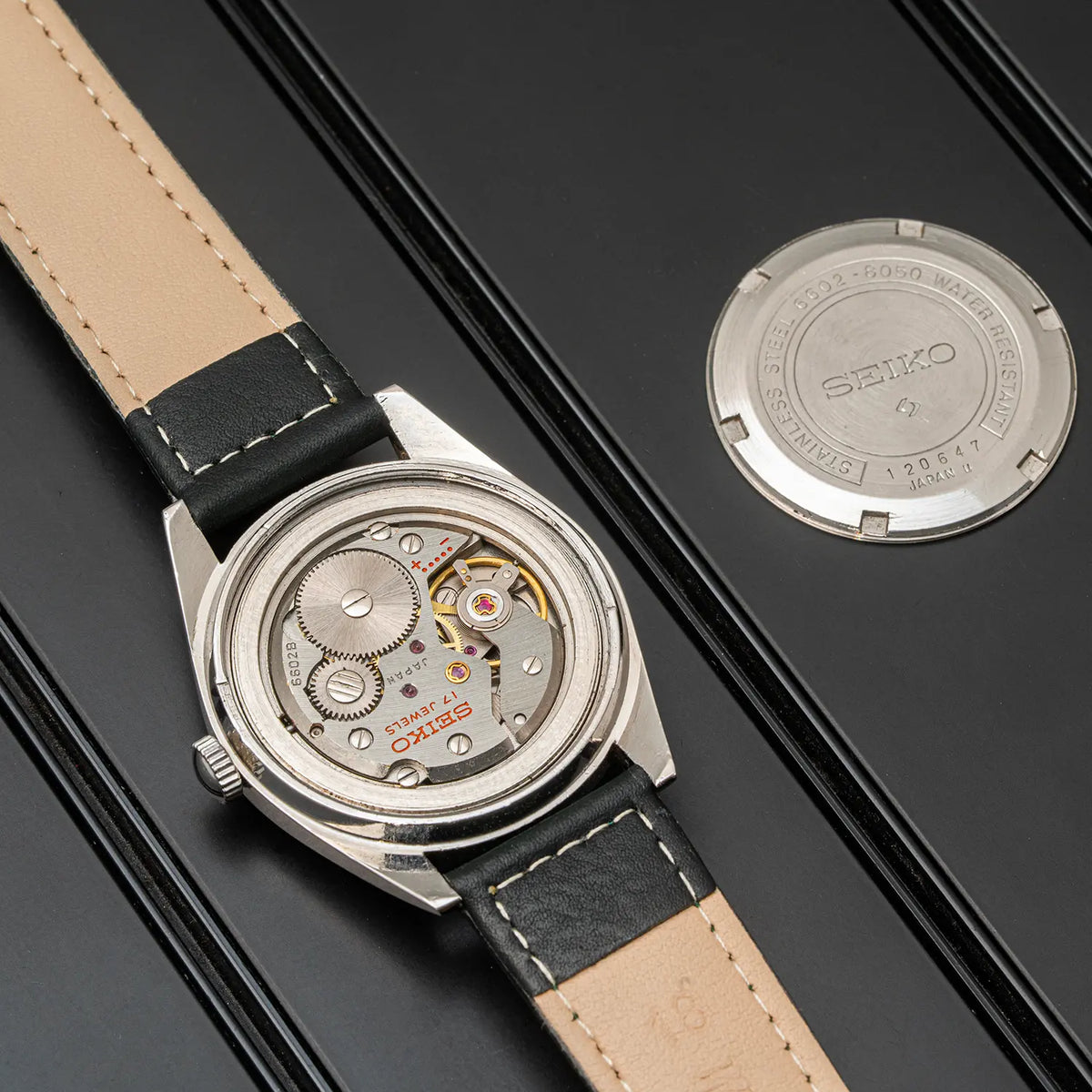 A Seiko, Ref. 6602-8050, Cal. 6602, Men’s Manual Wind, 1971 watch with a silver-toned case, beige leather strap, and open dial showing the mechanical movement, displayed on a black surface with the watch back shown separately.