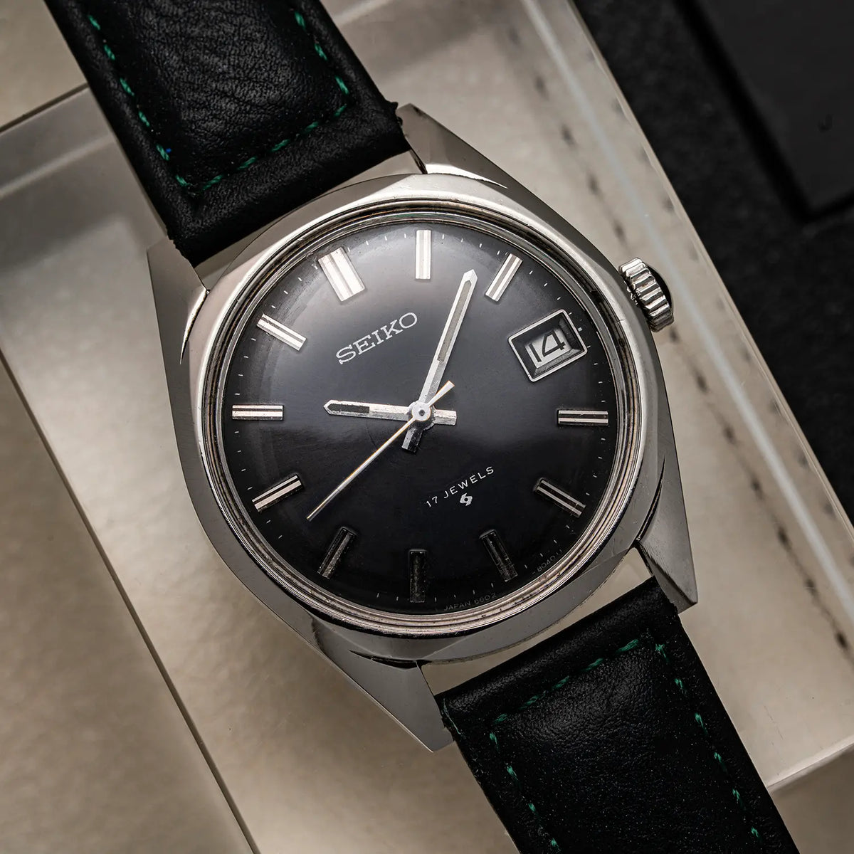 A vintage Seiko, Ref. 6602-8050, Cal. 6602, Men’s Manual Wind, 1971wristwatch with a black dial, silver-toned case, and black leather strap with green stitching, resting on a light-colored surface.