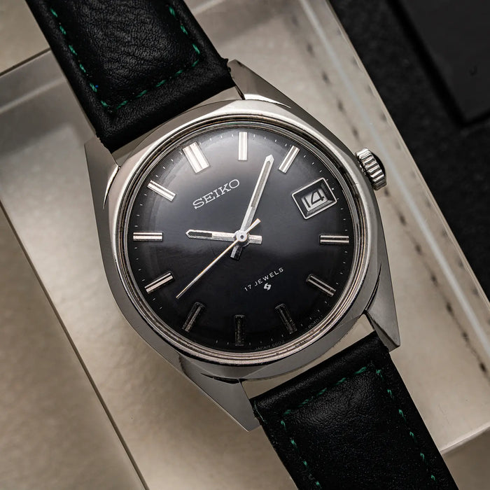 A vintage Seiko, Ref. 6602-8050, Cal. 6602, Men’s Manual Wind, 1971wristwatch with a black dial, silver-toned case, and black leather strap with green stitching, resting on a light-colored surface.