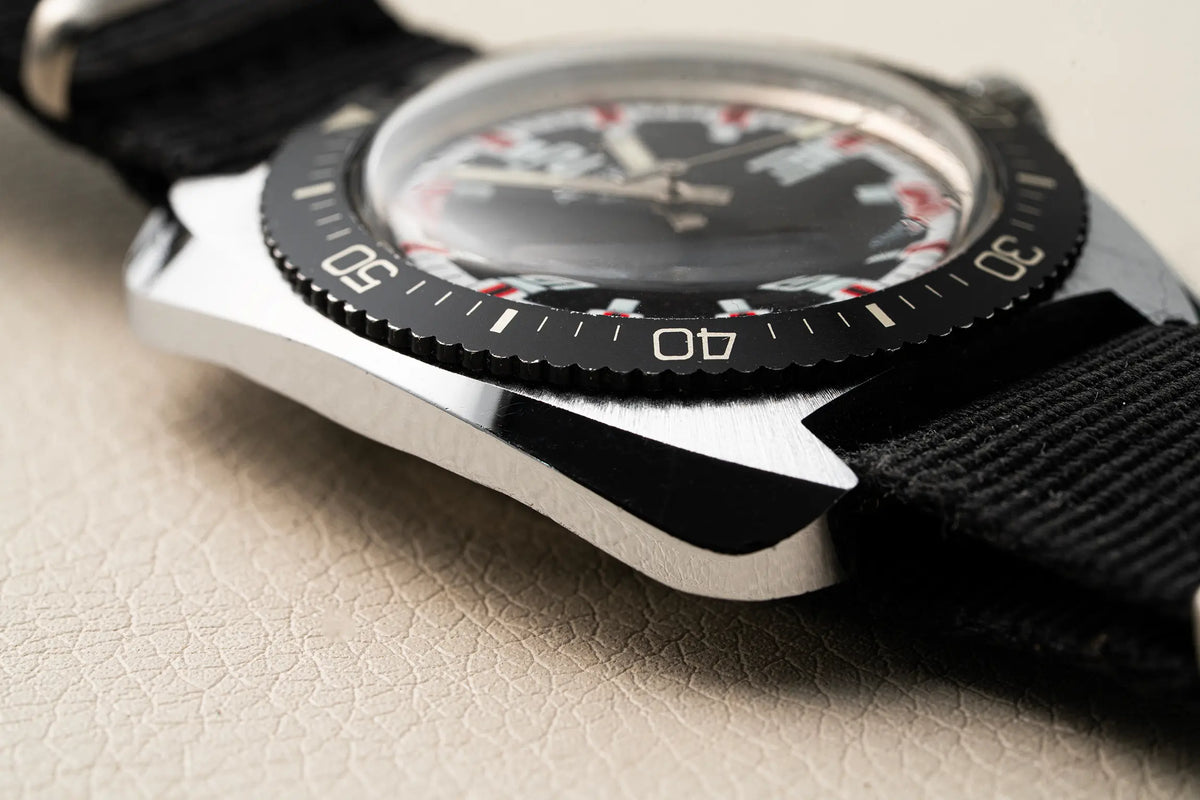 Close-up of a wristwatch Anker KR Diver, Cal. UMF 24, Men’s Diving Vintage Watch with a silver-toned metal case, black dial, black bezel, and black fabric strap on a light beige textured surface.