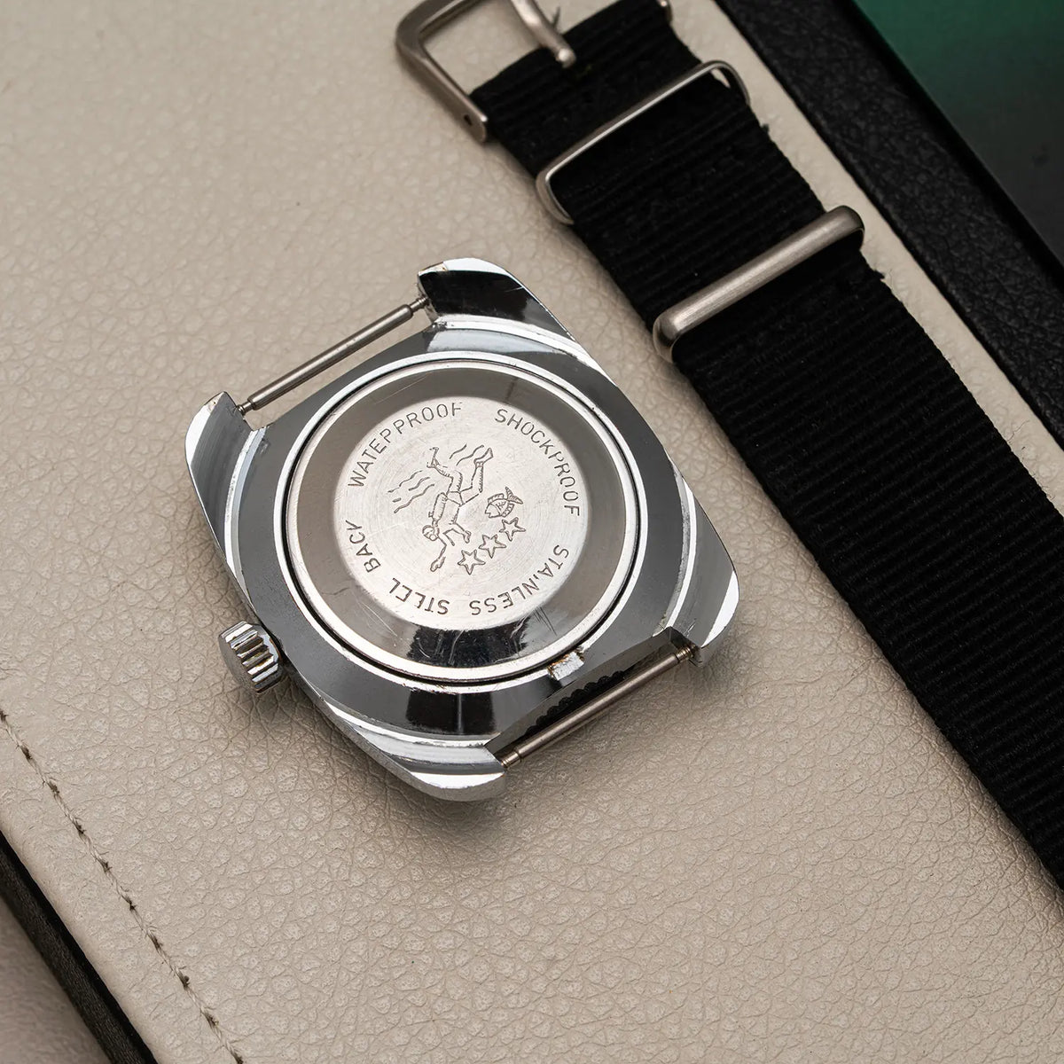 Close-up of the back of a stainless steel Anker KR Diver, Cal. UMF 24, Men’s Diving Vintage Watchwristwatch with a black fabric strap, placed on a light beige textured surface.