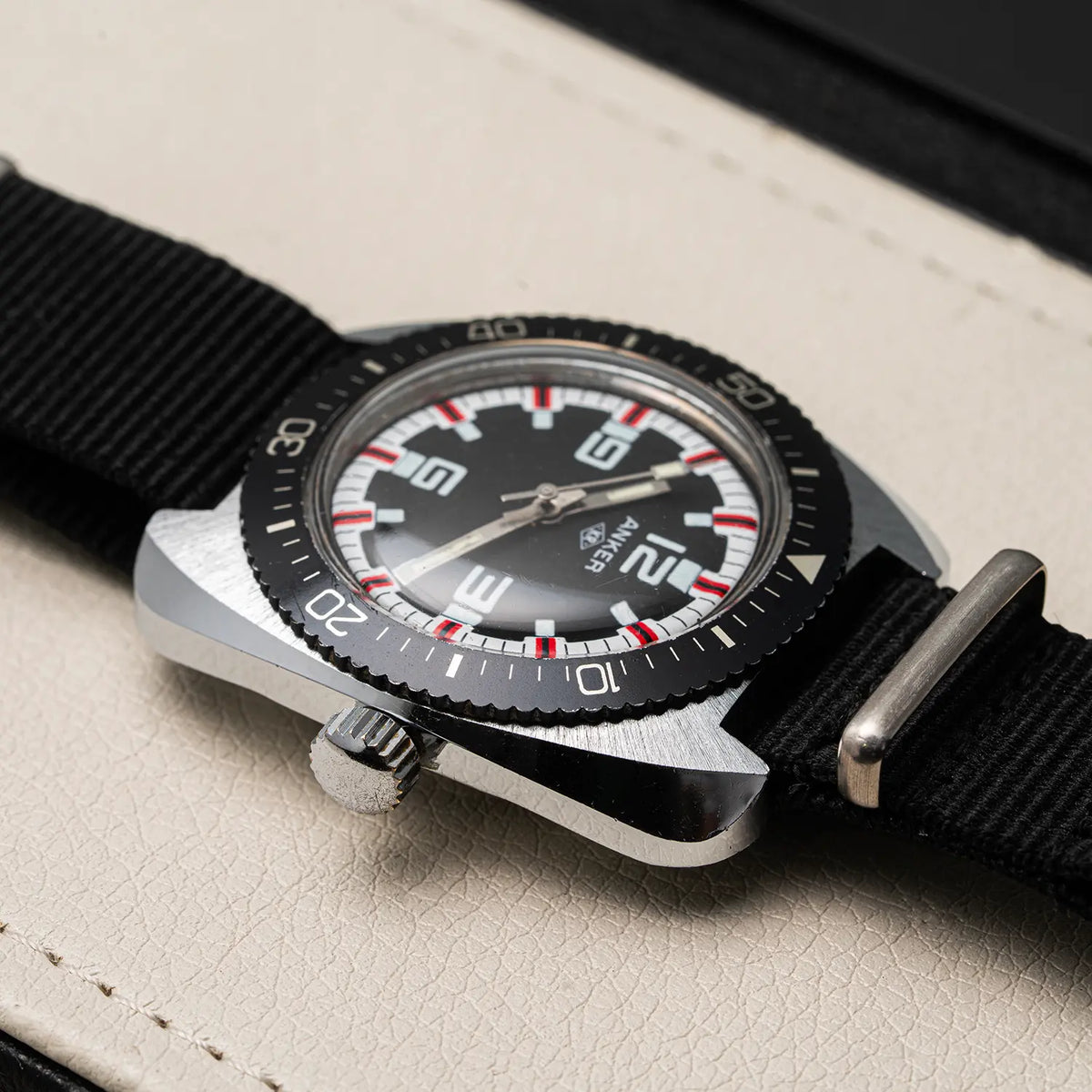 A close-up of a wristwatch Anker KR Diver, Cal. UMF 24, Men’s Diving Vintage Watch with a black dial, black rotating bezel, silver-toned metal case, and black fabric strap, resting on a light textured surface.