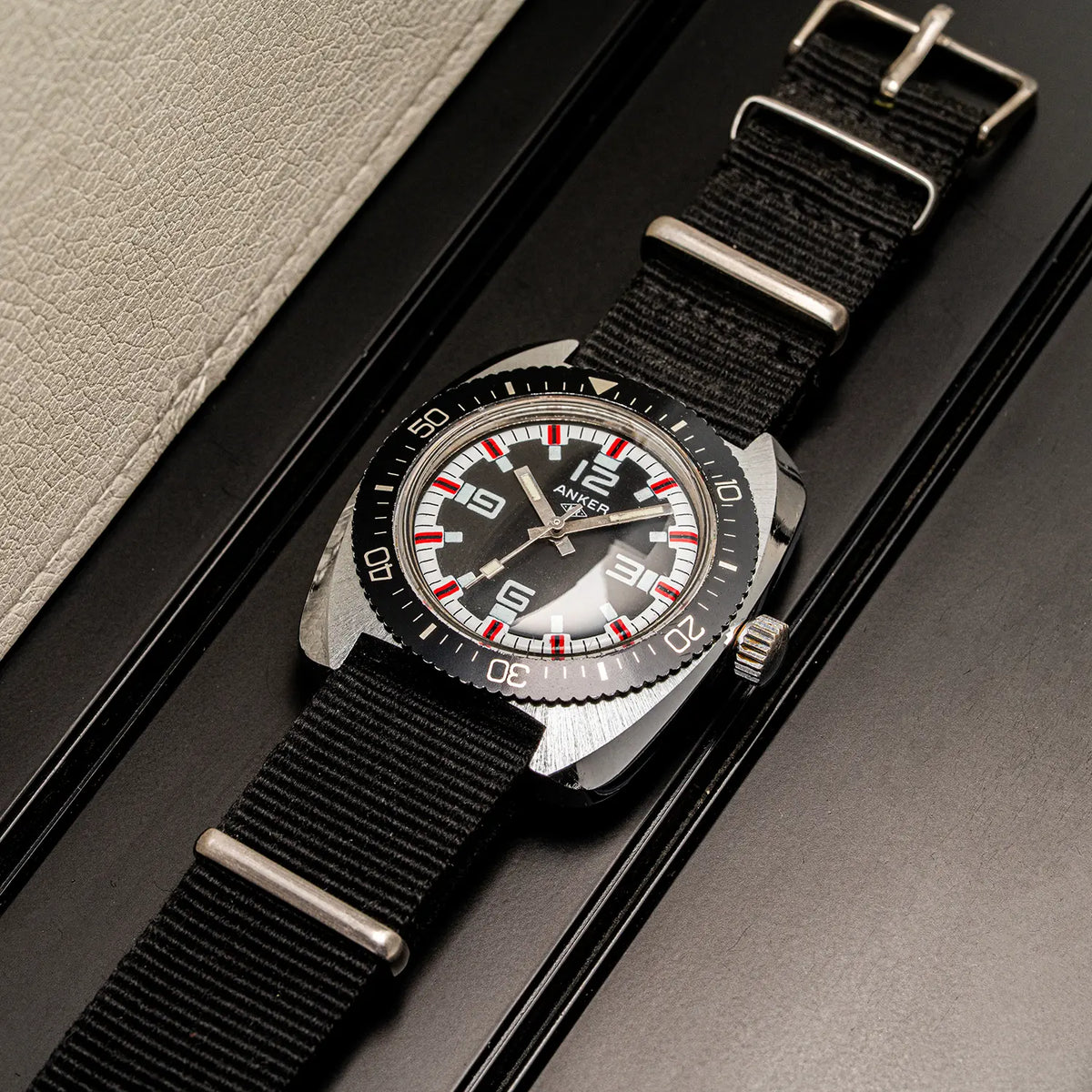 A wristwatch Anker KR Diver, Cal. UMF 24, Men’s Diving Vintage Watch with a black dial, silver-tone case, and black nylon strap, photographed diagonally on a dark surface next to a light beige textured surface.
