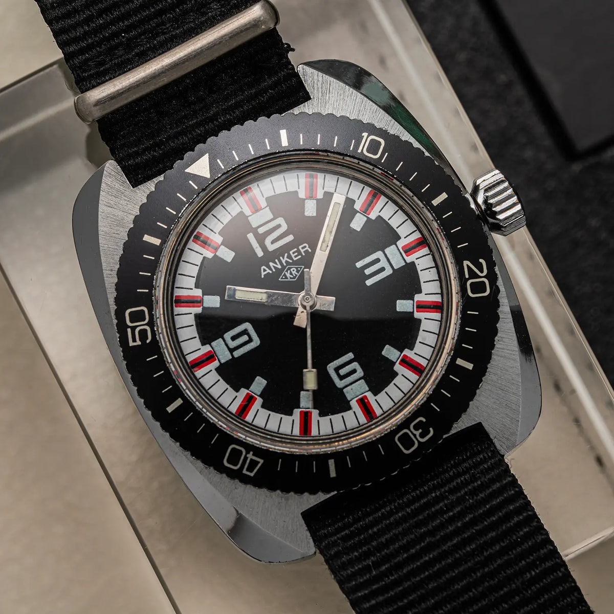 An Anker KR Diver, Cal. UMF 24, Men’s Diving Vintage Watchwristwatch with a square-shaped brushed steel case, black dial, black bezel with white and red markings, and a black fabric strap, shown resting on a light-colored surface.