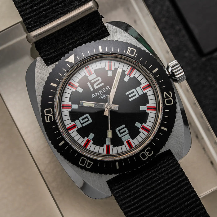 An Anker KR Diver, Cal. UMF 24, Men’s Diving Vintage Watchwristwatch with a square-shaped brushed steel case, black dial, black bezel with white and red markings, and a black fabric strap, shown resting on a light-colored surface.
