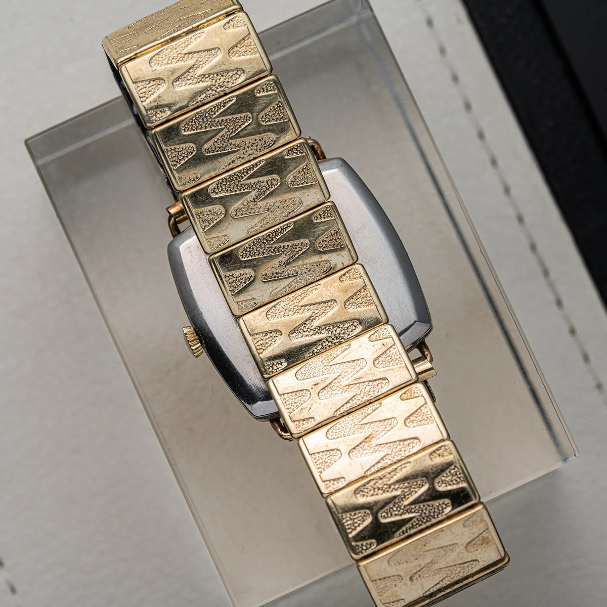 A close-up of a vintage gold-tone watch Bergana PUW 460 with a distinctive textured link bracelet, resting on a clear display stand.
