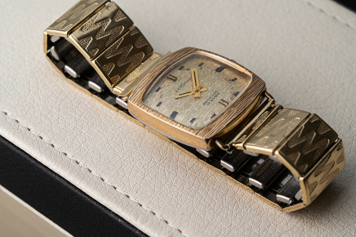 A vintage gold-tone wristwatch Bergana PUW 460 with a textured chevron-patterned metal bracelet and a square dial, resting on a light beige textured leather surface.