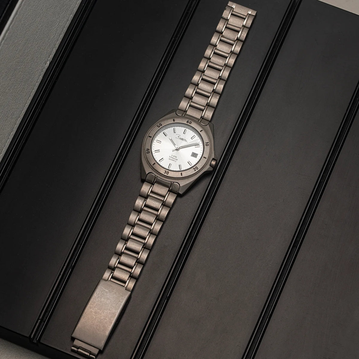A vintage-style Bossini Titanium wristwatch with a silver-toned metal link bracelet and a white dial, displayed diagonally on a dark textured surface.