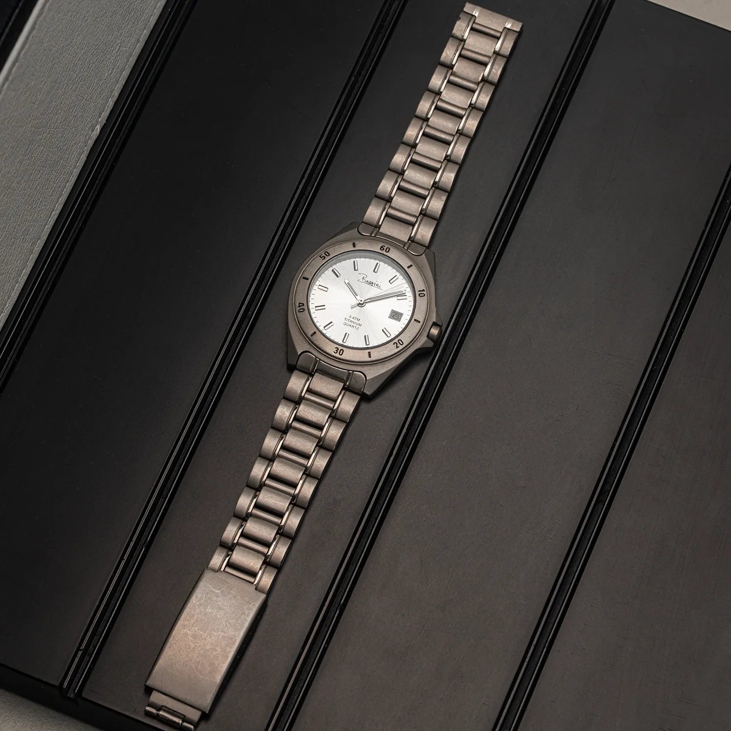 A vintage-style Bossini Titanium wristwatch with a silver-toned metal link bracelet and a white dial, displayed diagonally on a dark textured surface.