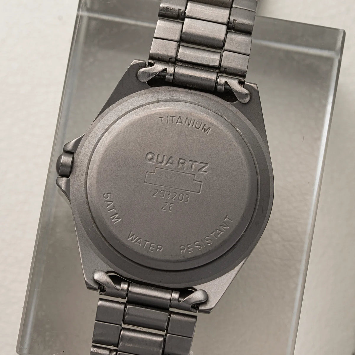 The back of a Bossini Titanium quartz watch with a metal link bracelet, resting on a clear acrylic stand. The case is engraved with 'TITANIUM', 'QUARTZ', and 'SWIM WATER RESISTANT'.