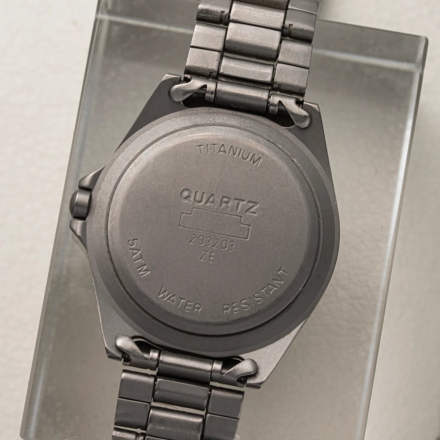 The back of a Bossini Titanium quartz watch with a metal link bracelet, resting on a clear acrylic stand. The case is engraved with 'TITANIUM', 'QUARTZ', and 'SWIM WATER RESISTANT'.