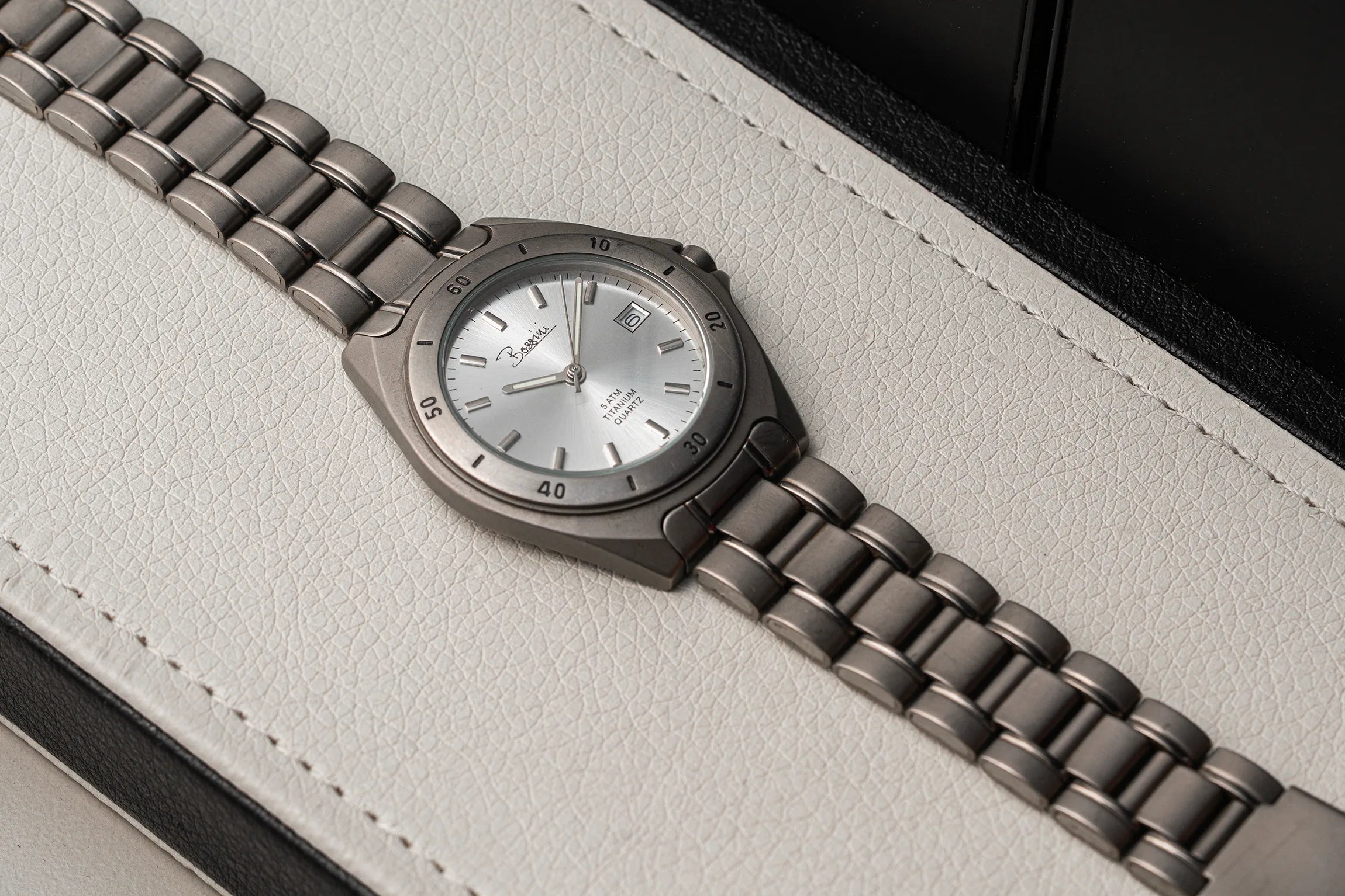 A close-up of a wristwatch Bossini Titanium with a dark gunmetal gray metal link bracelet and a silver dial, resting on a textured light gray leather surface.