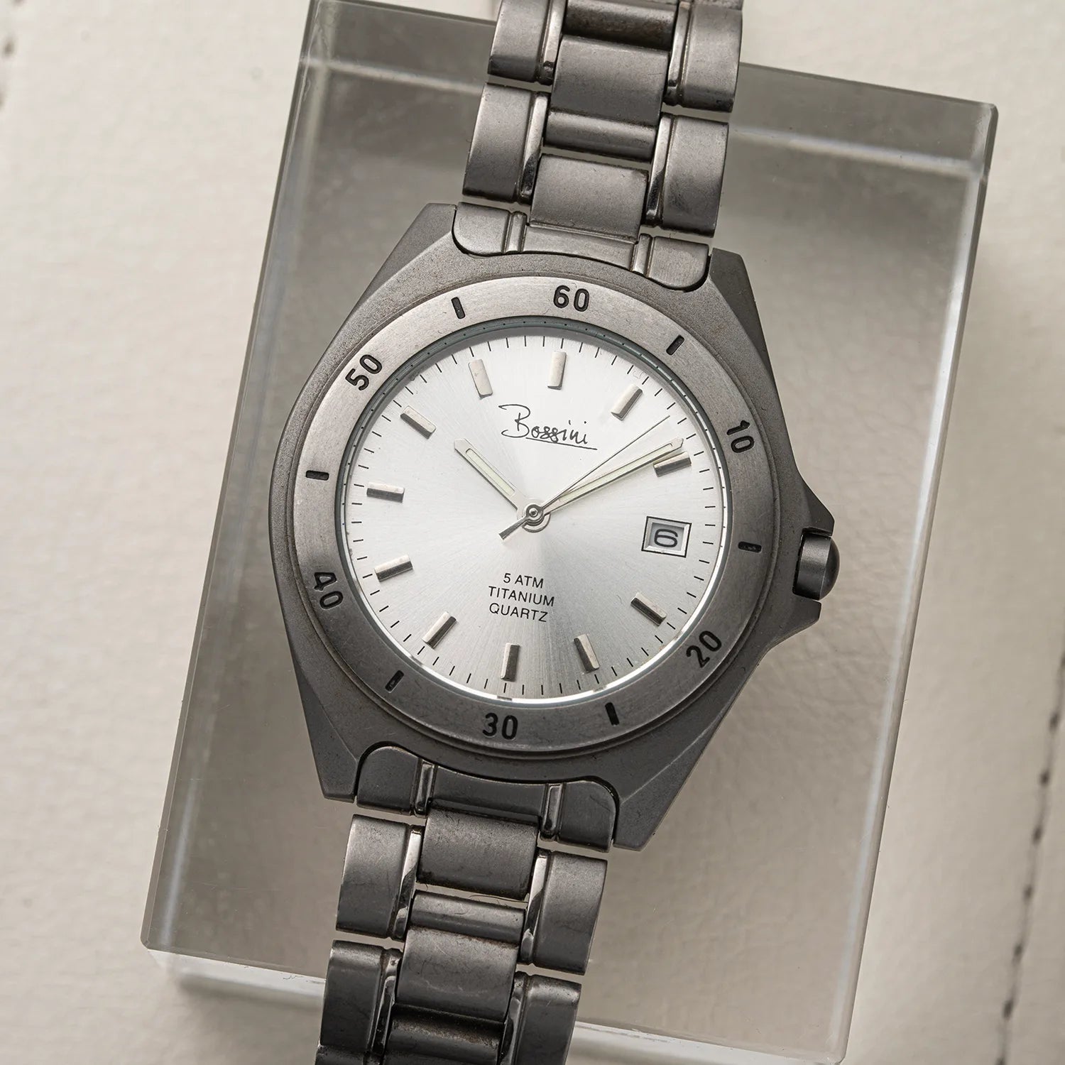 A Barretti titanium watch Bossini Titanium with a dark gray metal link bracelet and a silver dial, displayed on a clear acrylic stand.