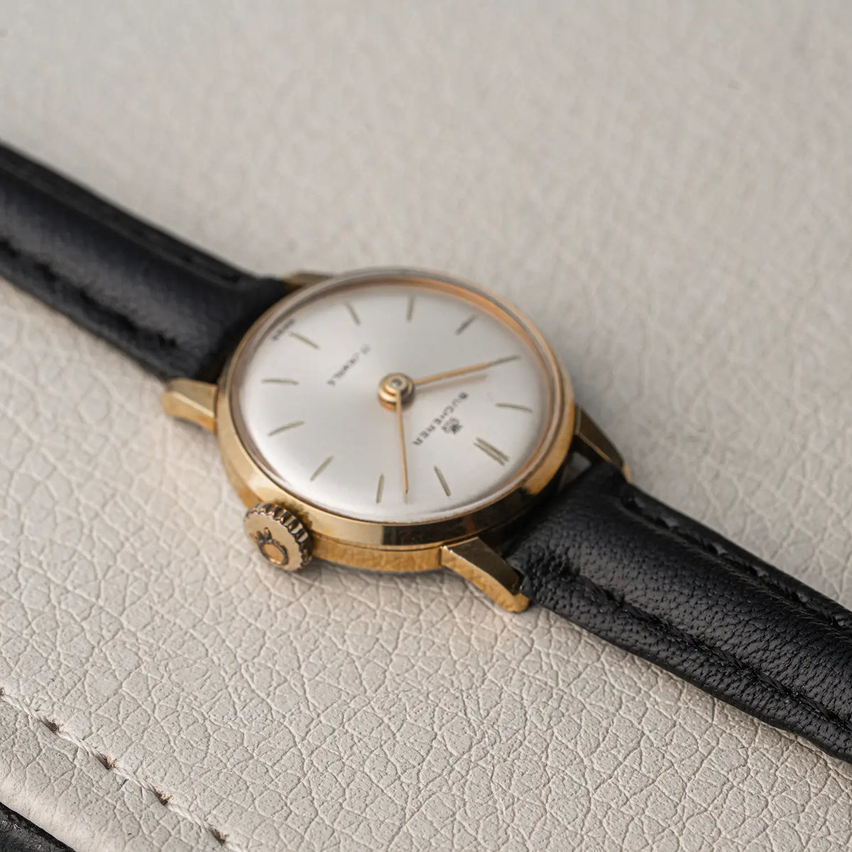 A vintage wristwatch with a gold-toned case, white dial, and black leather strap, resting on a textured light beige surface.