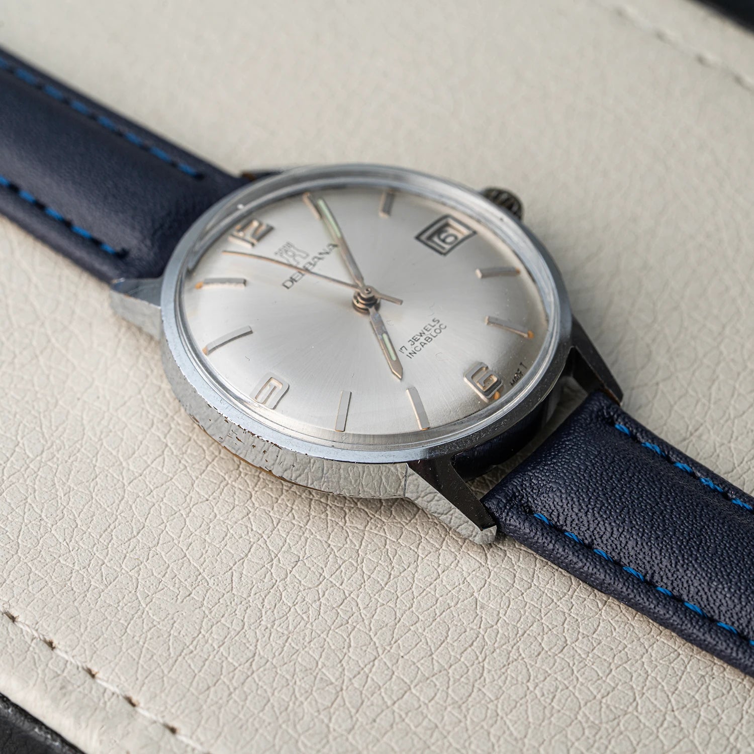A vintage-style wristwatch Delbana, Cal. AS 1951 with a silver-toned round case, silver dial, date window, and dark blue leather strap with light blue stitching, photographed close up on a cream textured surface.