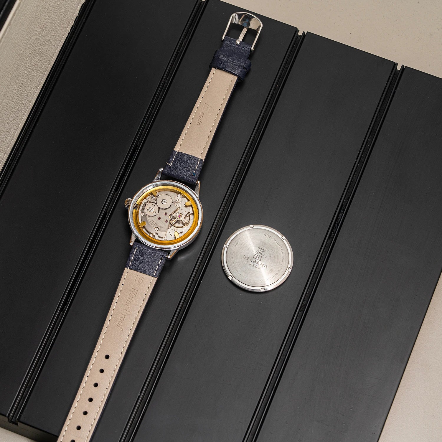 Cal. AS 1951  with a beige leather strap and navy blue accents, displayed diagonally on a black presentation box next to its silver metal back cover.