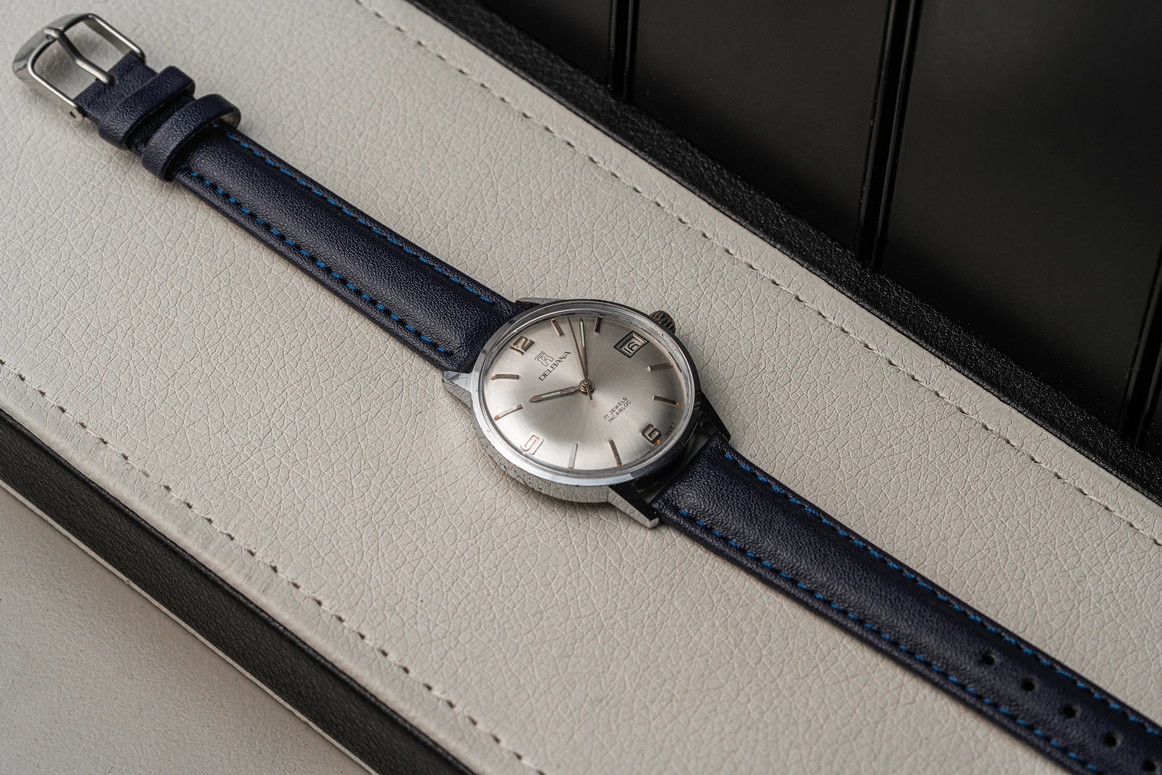 A vintage-style wristwatch Delbana, Cal. AS 1951 with a silver dial and a dark blue leather strap with blue stitching, resting diagonally on a light beige textured leather surface.